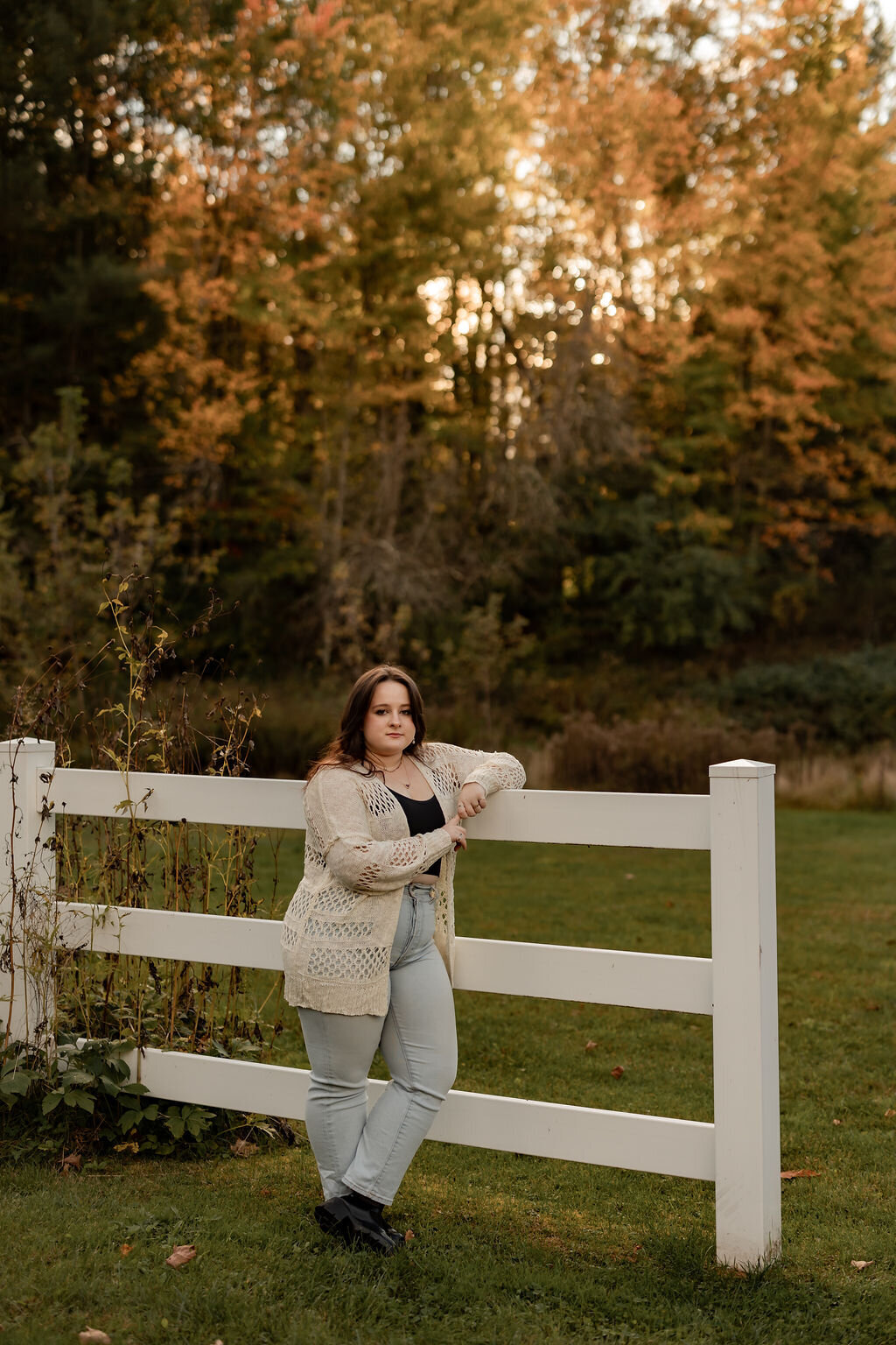 A person with long hair stands outdoors leaning on a white fence, perfectly capturing the essence of a New England photo book. They wear a light-colored knit cardigan over a dark top and light jeans. Autumn trees with vibrant leaves fill the background, and the grass is lush and green.
