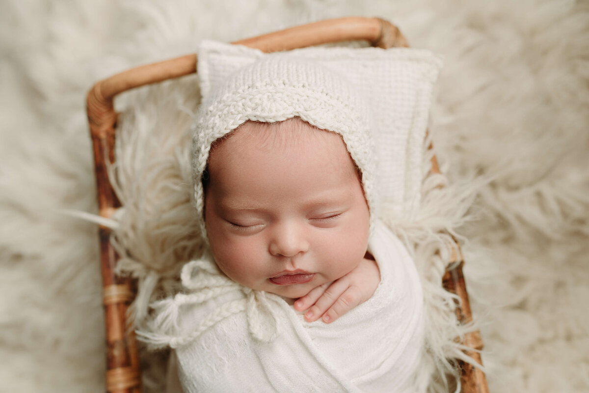 A newborn baby sleeps in a white knit bonnet in a small wicker bed