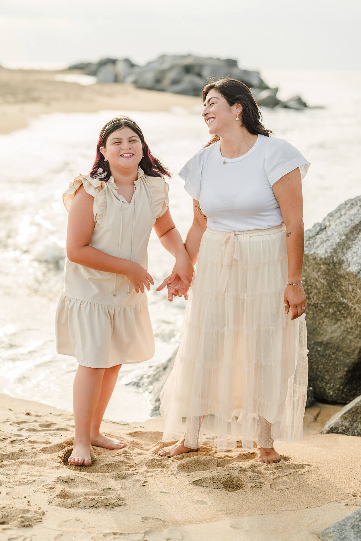 A mother, wearing an off-white skirt and white top, holds her daughter's hand, The daughter, in an off-white dress, laughs with her mom.