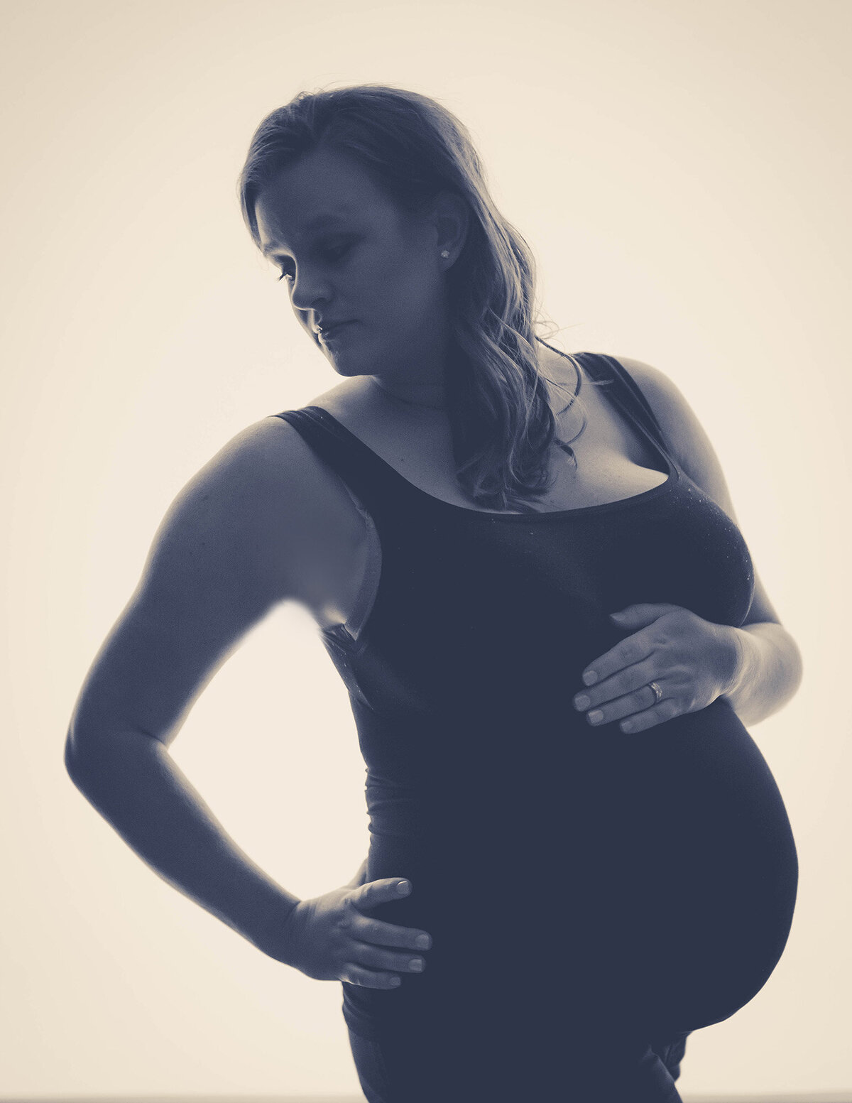 A mother to be stands in a studio looking over her shoulder with a hand on her bump in a black maternity gown