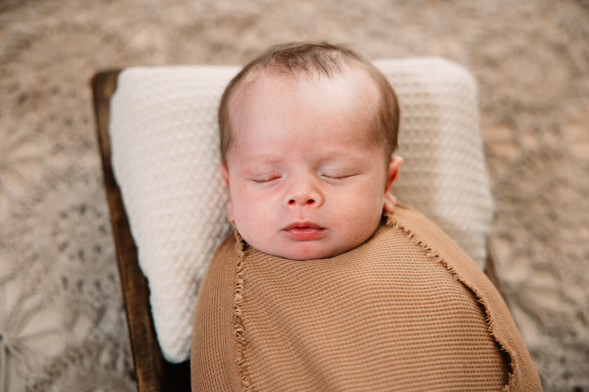 A newborn sleeps on a pillow in a brown swaddle