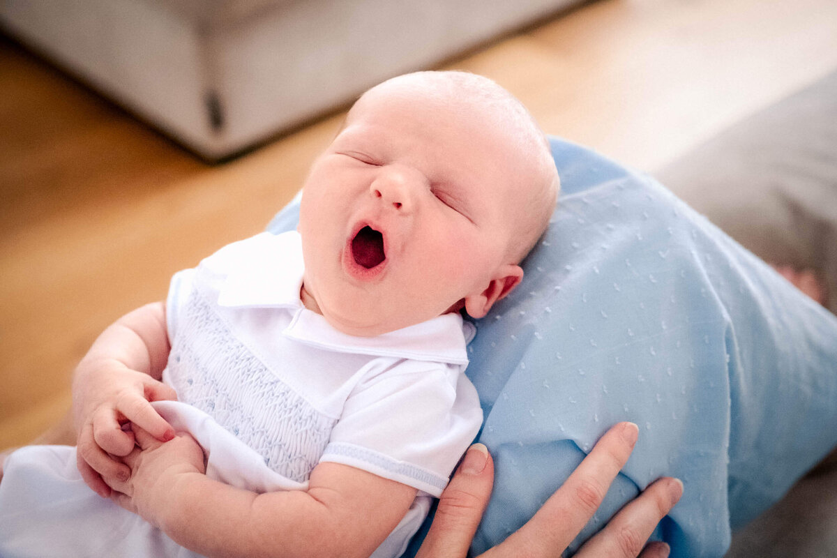 Newborn baby yawning while being held on top of lap covered in blue dress.