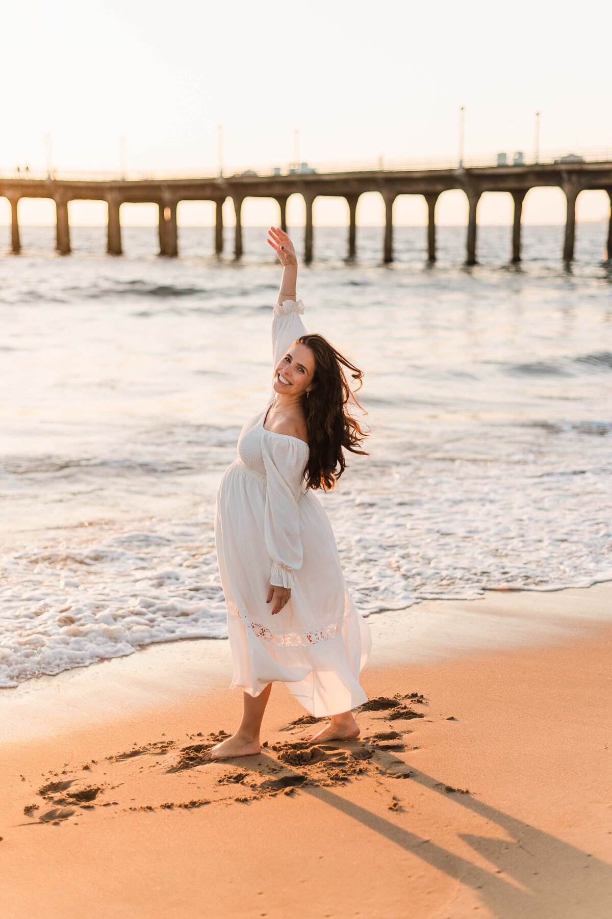 A pregnant woman smiling and dancing on the beach at sunset.