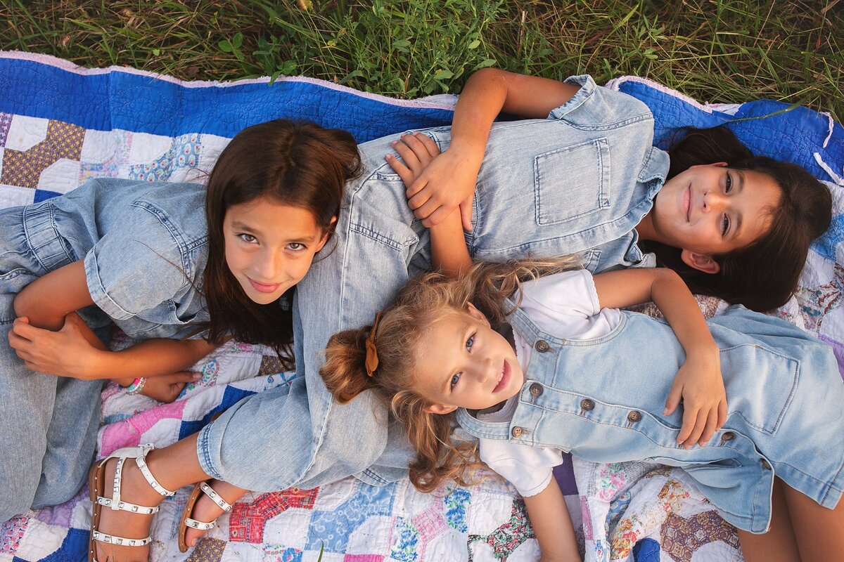 three-sisters-in-matching-denim-outfits-laying-on-colorful-quilt-outdoors-smiling-during-family-photography-session-in-fairland