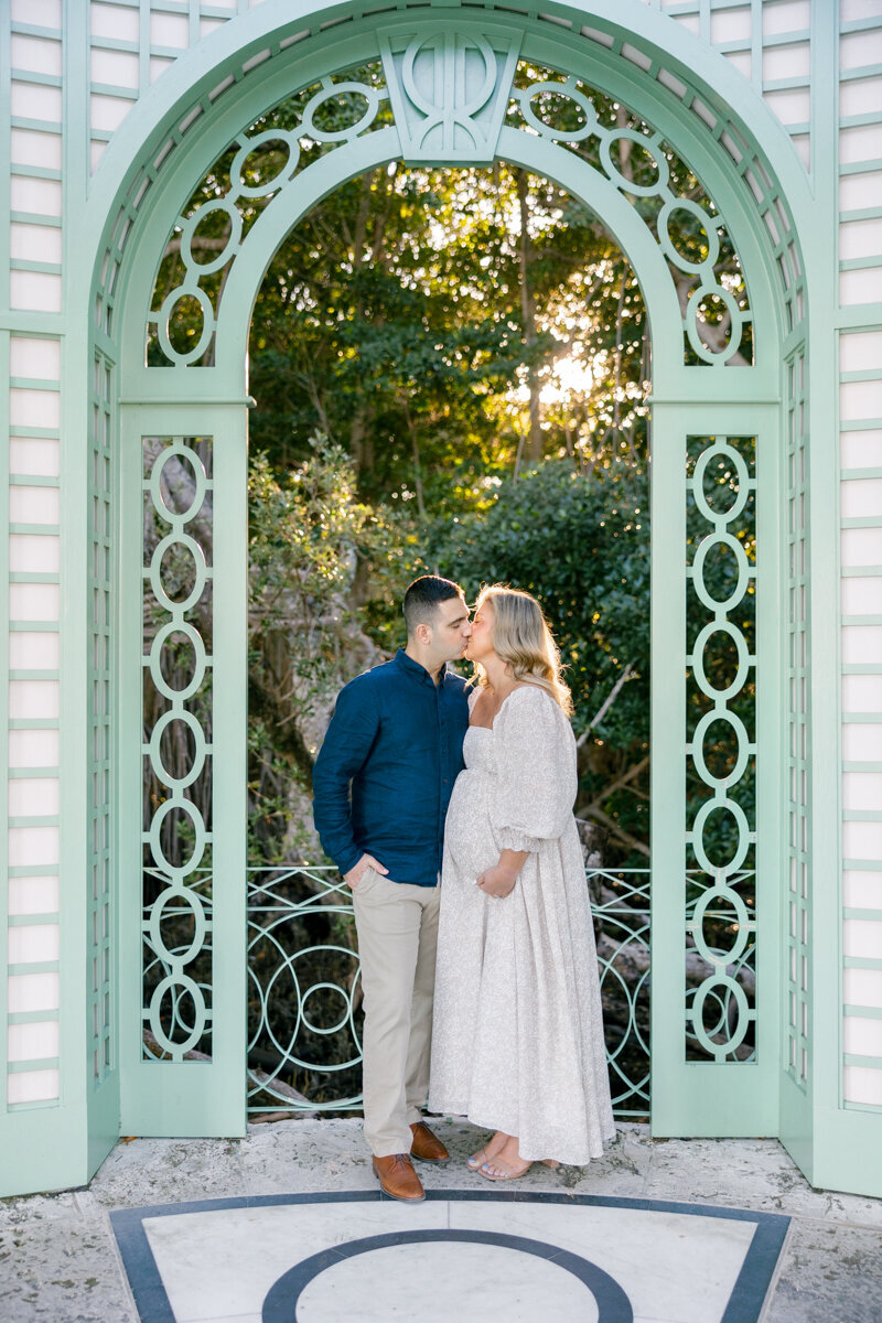 A couple shares a loving moment beneath a pastel-green arched doorway during their Florida maternity session.
