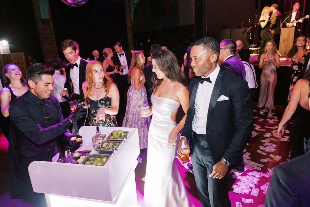 The newlyweds walk through a lively reception space, illuminated with vibrant pink lighting, surrounded by guests.