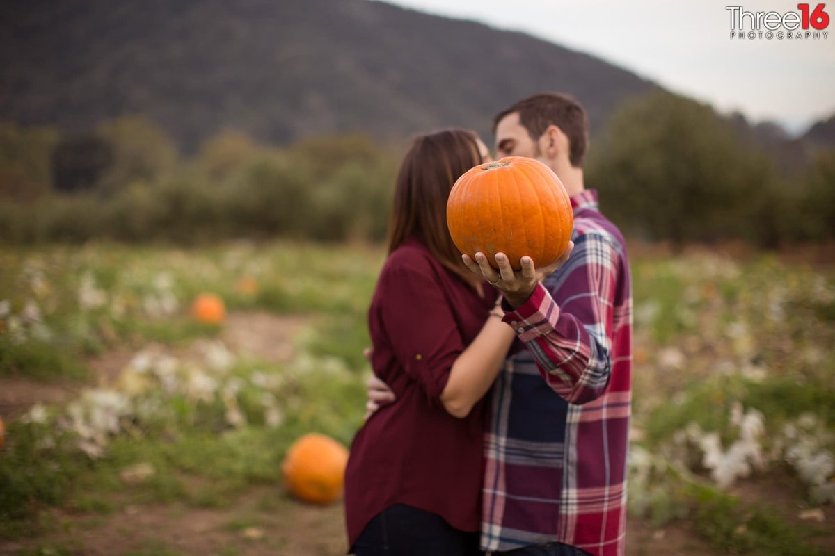 Engaged couple share a kiss behind a pumpkin while standing in a pumpkin patch