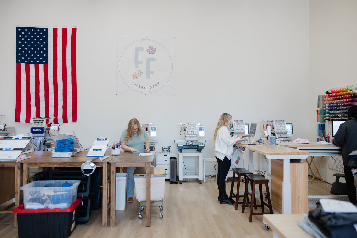Two women working at embroidery machines inside Floss and Fable’s bright studio space with FF logo on the wall. Photograph by Yucaipa branding photographer Kaitlyn Dawn Photography.