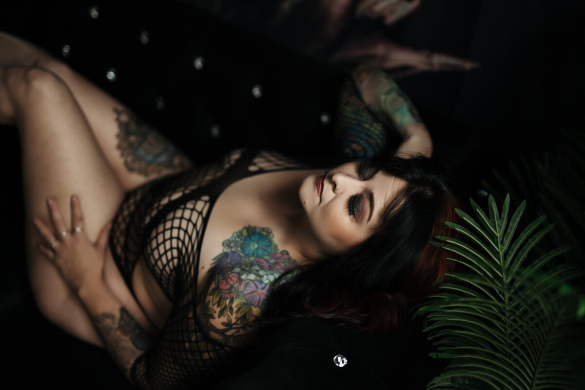 A woman with tattoos leans back on a blue couch under a window  while wearing black mesh lingerie