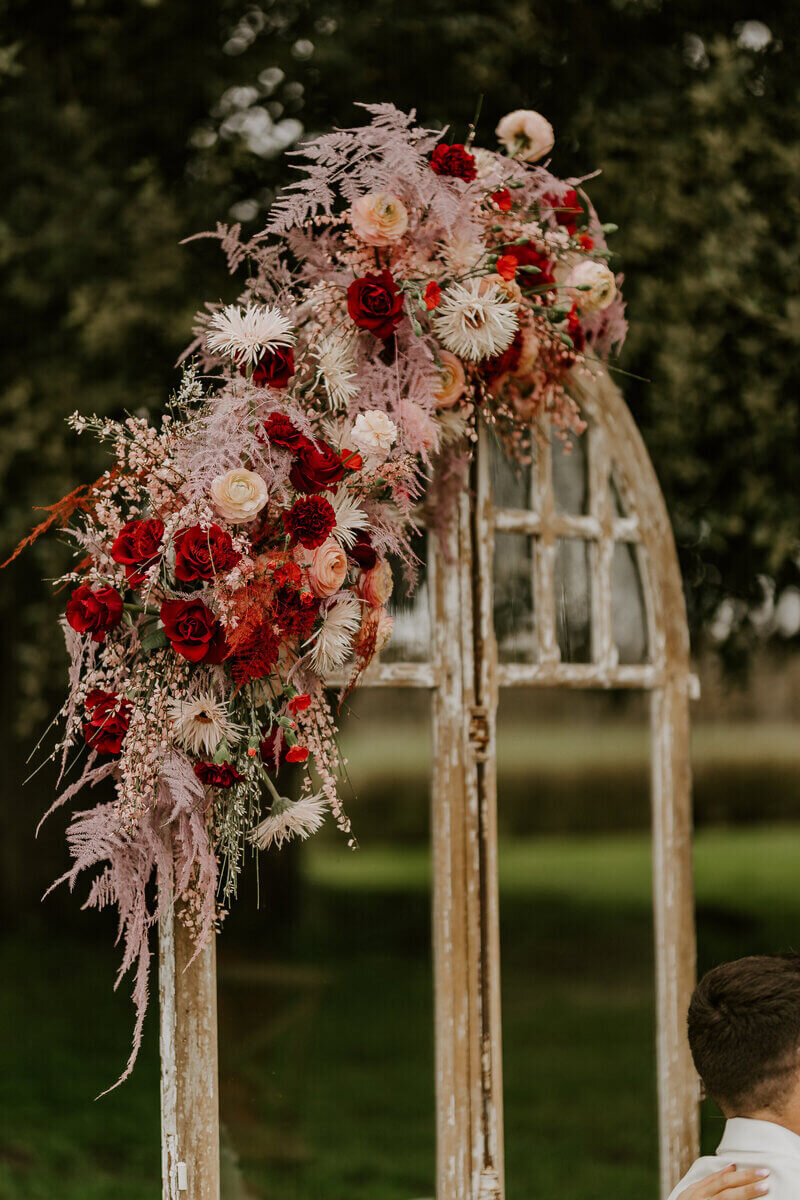 Composition florale rouge et rose surmontant une arche en bois dans un décor verdoyant. Photo prise par Laura Termeau photographie.