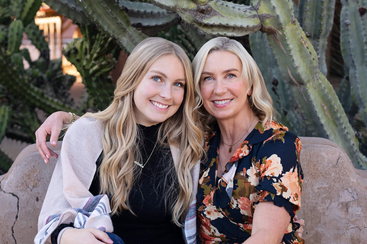A mother and daughter with blonde hair with their heads together smiling at the camera with greenery behind them.