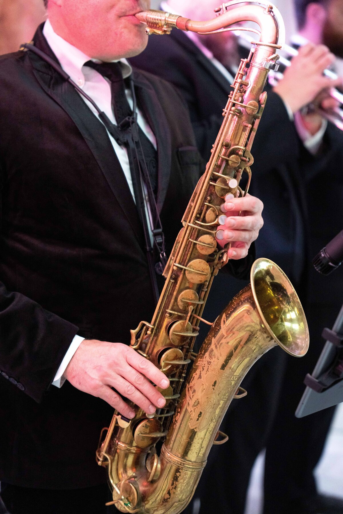 A musician in a suit plays a golden saxophone, focusing on the instruments keys. In the blurred background, another musician plays a trumpet. The scene conveys a live jazz or band performance.