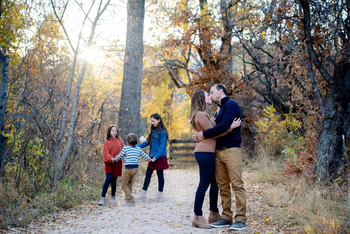 A mom and dad kiss in a park trail while their three young children dance behind them