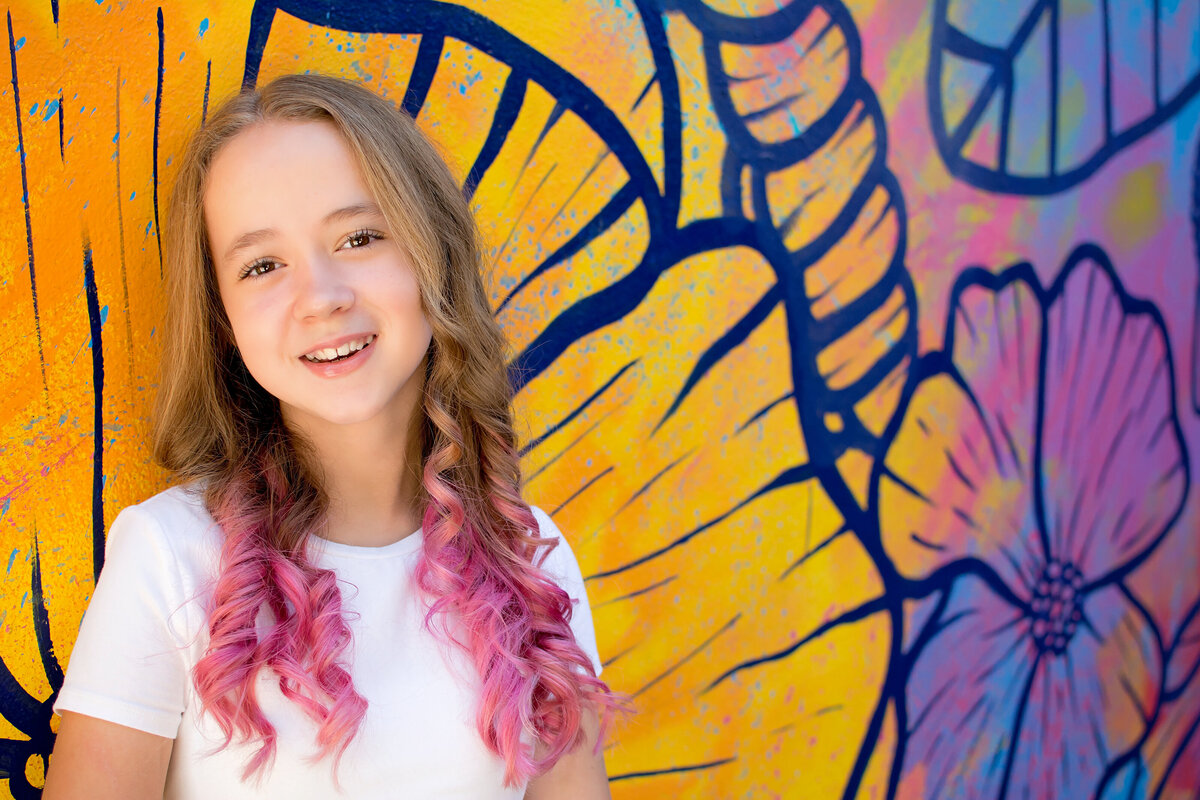 Tween girl standing in front of floral painted wall with white shirt and pink hair