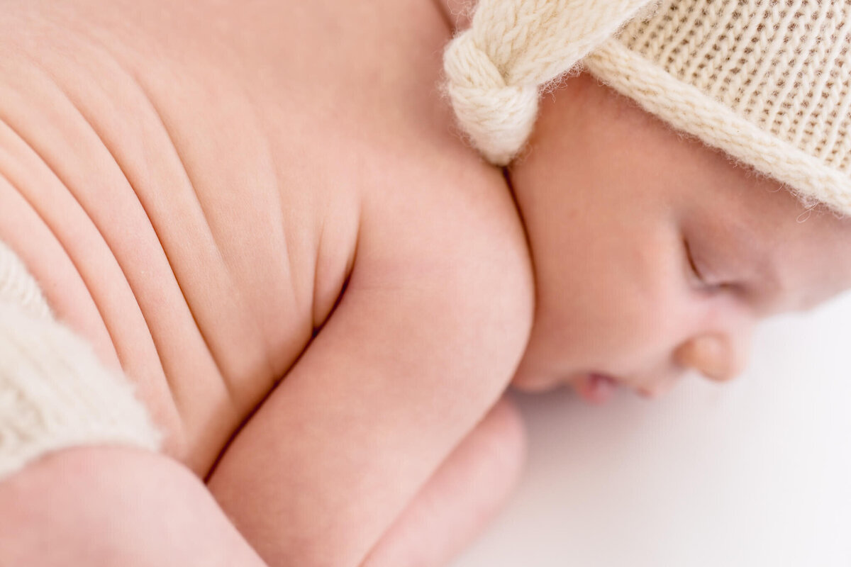 Close-up of a sleeping baby lying on their stomach, wearing a knitted cream-colored hat and matching diaper cover. The baby has soft, smooth skin with gentle rolls. The background is a soft white, enhancing the peaceful, cozy atmosphere.