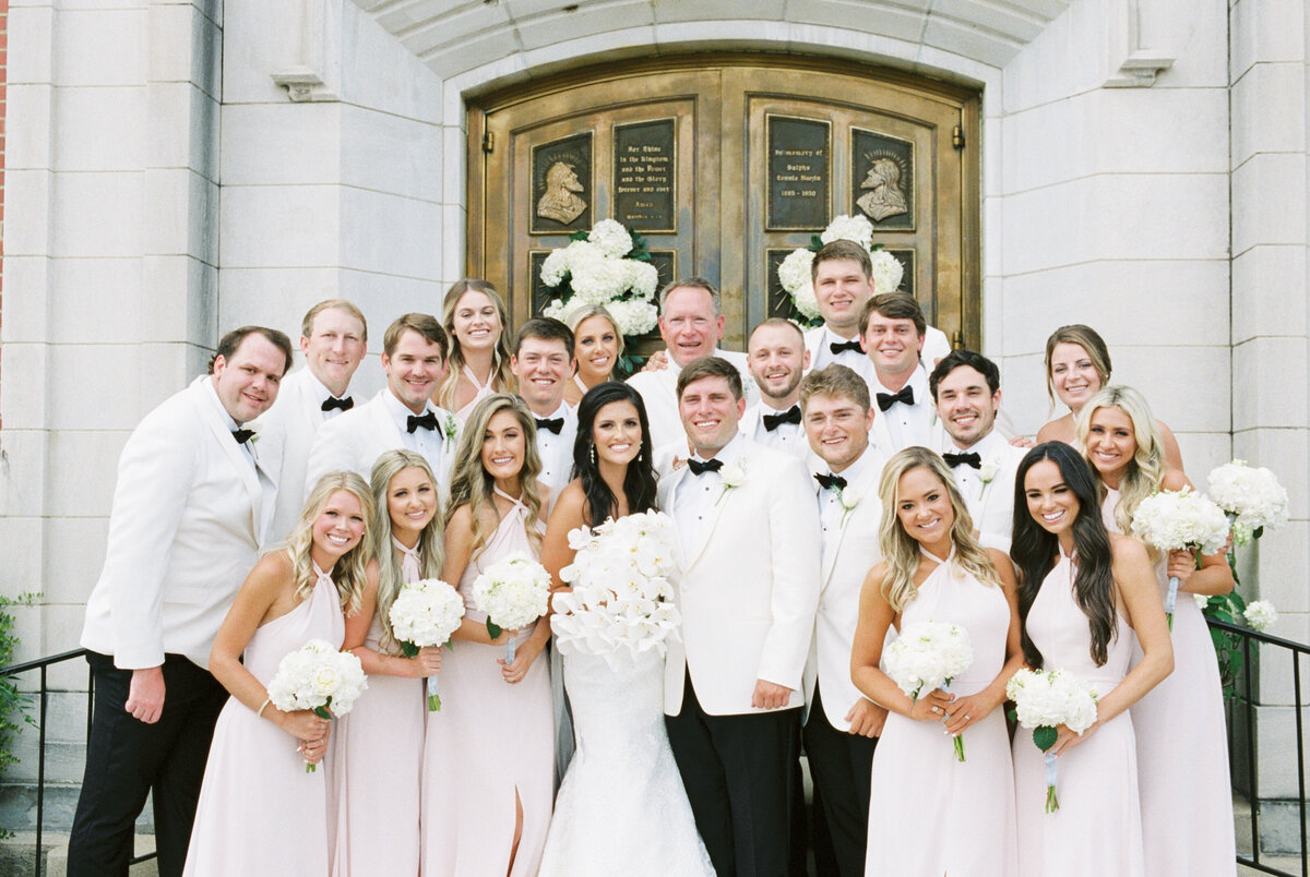 A wedding party stands to smile in front of beautiful antique church doors by Birmingham wedding photographer, Kelsey Dawn Photography