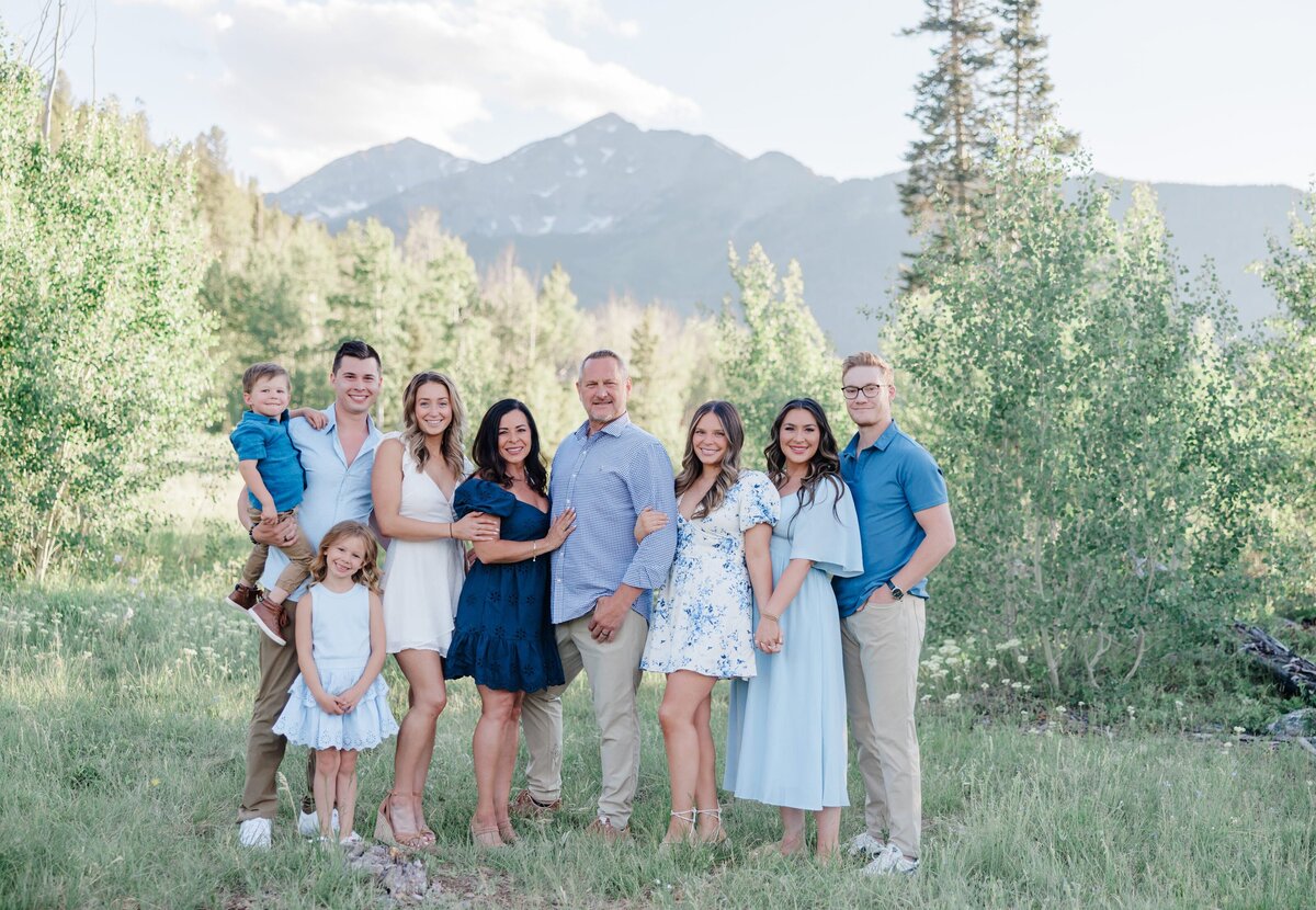 An extended family with 3 generations stands together with hands on each other in a forest in summer