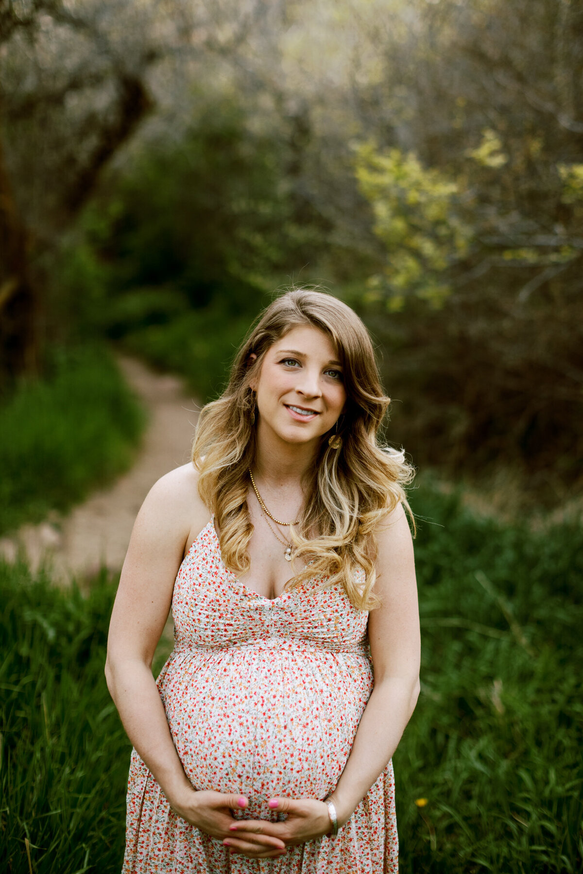 woman in a sun dress  holds her pregnant stomach near a trail in boulder, colorado