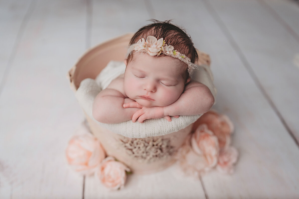 Sleeping newborn girl posing with head on hands sitting in a cream color bucket on white wood floor with pink flowers around it