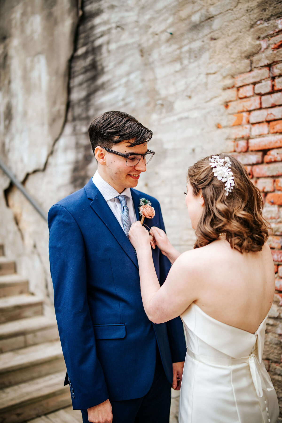 A bride in a strapless white dress pins a boutonniere on the smiling groom’s blue suit. They stand together by a weathered brick and plaster wall with wooden stairs beside them.