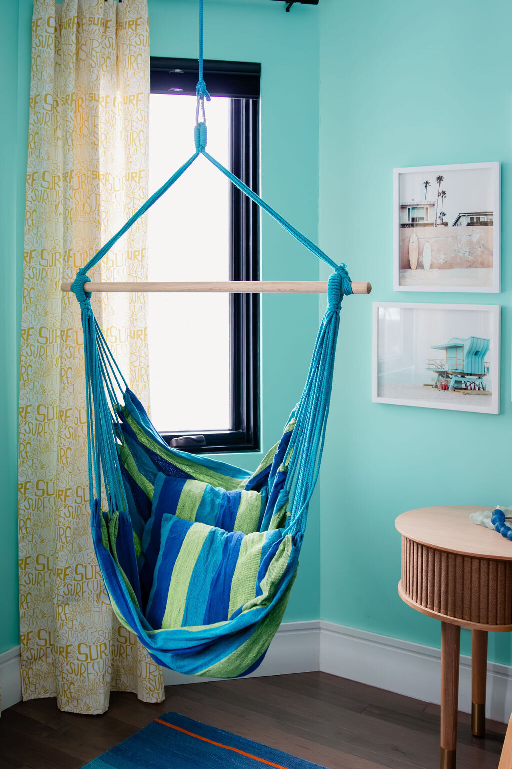A bright room with light turquoise walls features a blue and green striped hammock chair hanging from the ceiling near a window with patterned curtains. The hammock chair is supported by a wooden spreader bar and contains blue and green striped pillows. To the right, a light-colored Umage Audacious side table with tambour detailing and slender legs stands against the wall. Above the table, two framed prints with beach-themed imagery hang on the wall. A glimpse of a colorful striped rug is visible on the dark wooden floor below the hammock chair.