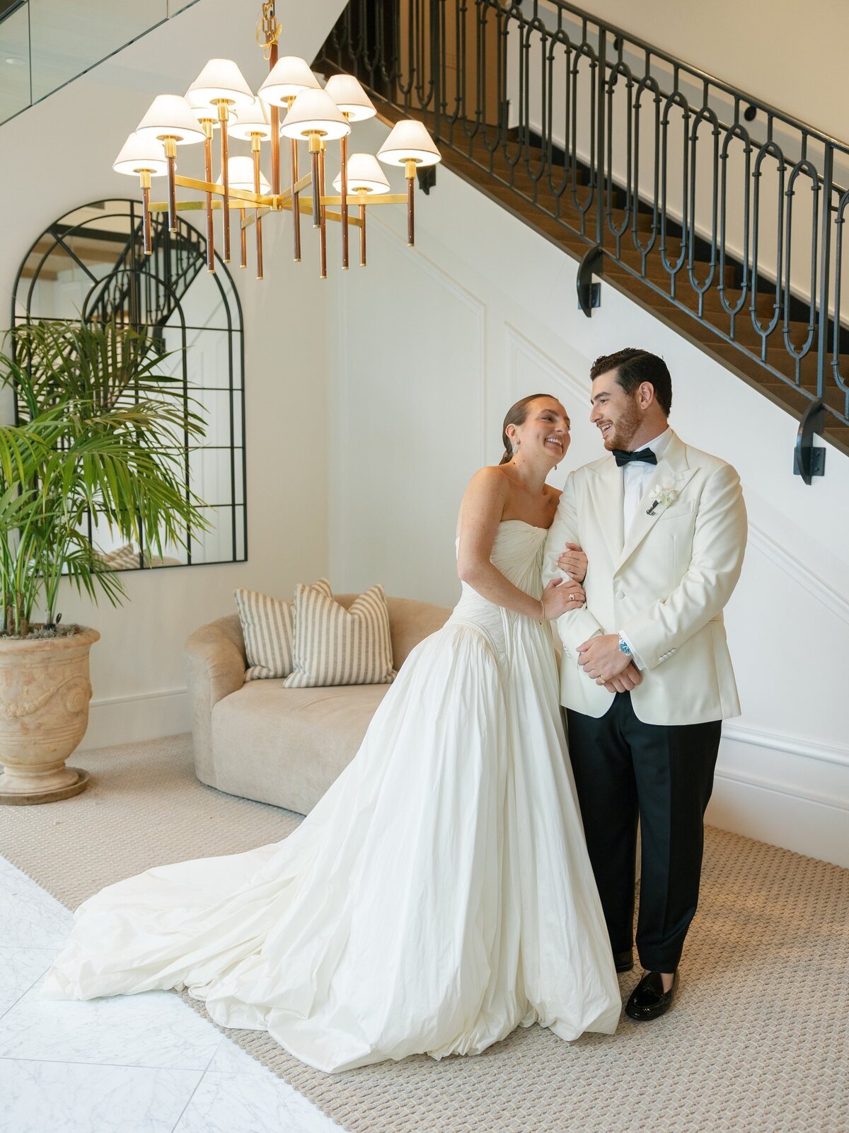 Kara and Nick smiling at each other beneath the staircase and chandelier at Pendry Natirar