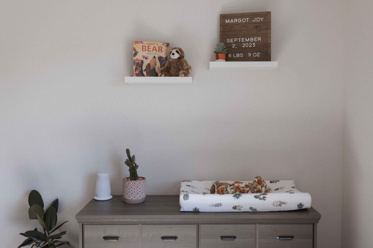 Minimalist baby room with a dresser, a cactus plant, and a changing table with a baby dressed in sunflower print. Wall shelves display a bear-themed book, a sloth stuffed toy, and a sign with the baby's birth details.