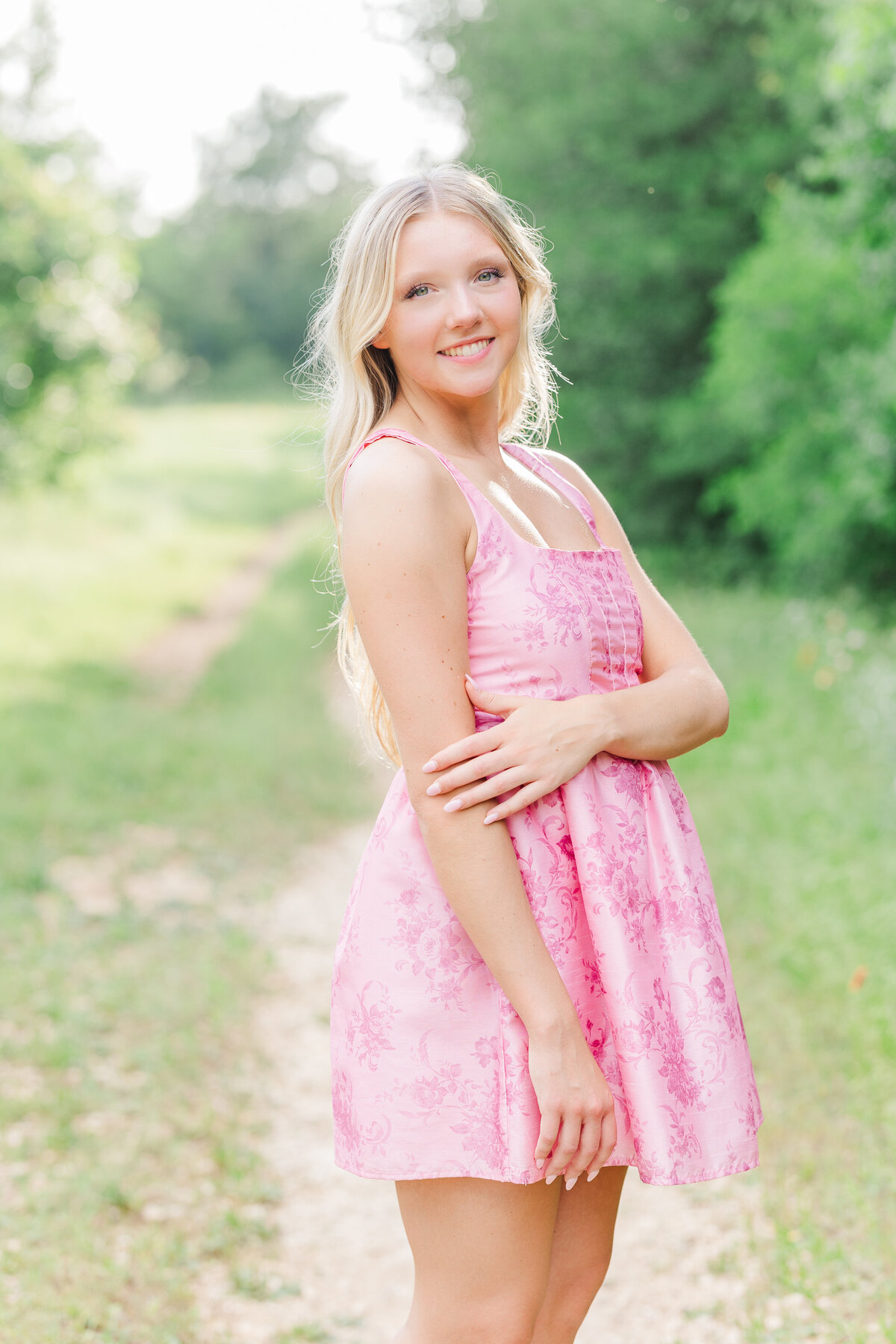 A joyful senior girl in a pink dress poses along a natural trail in Arbor Hills Nature Preserve, glowing in golden sunlight. Plano senior portraits that feel fresh, authentic, and effortless.