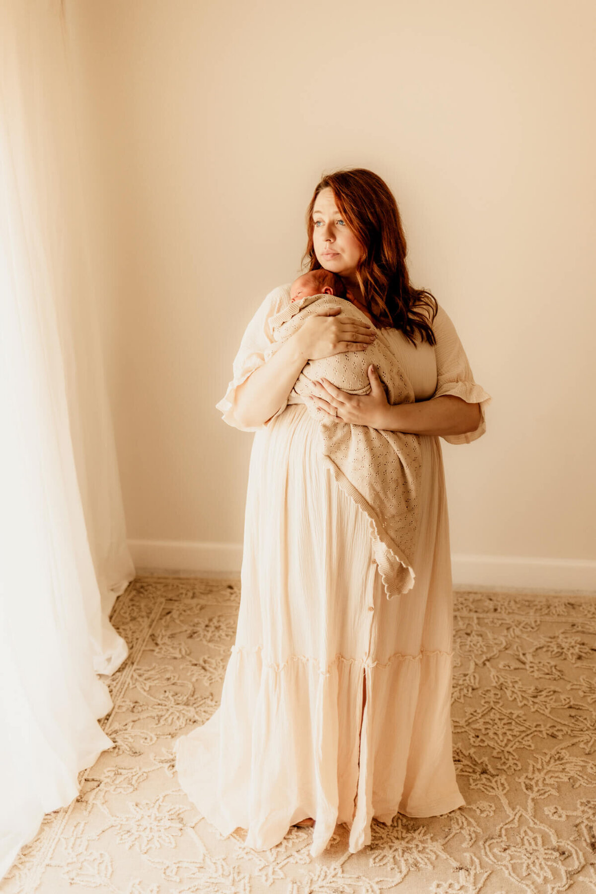 Mother with red hair wearing a cream dress holds her baby on her chest while looking out the window.