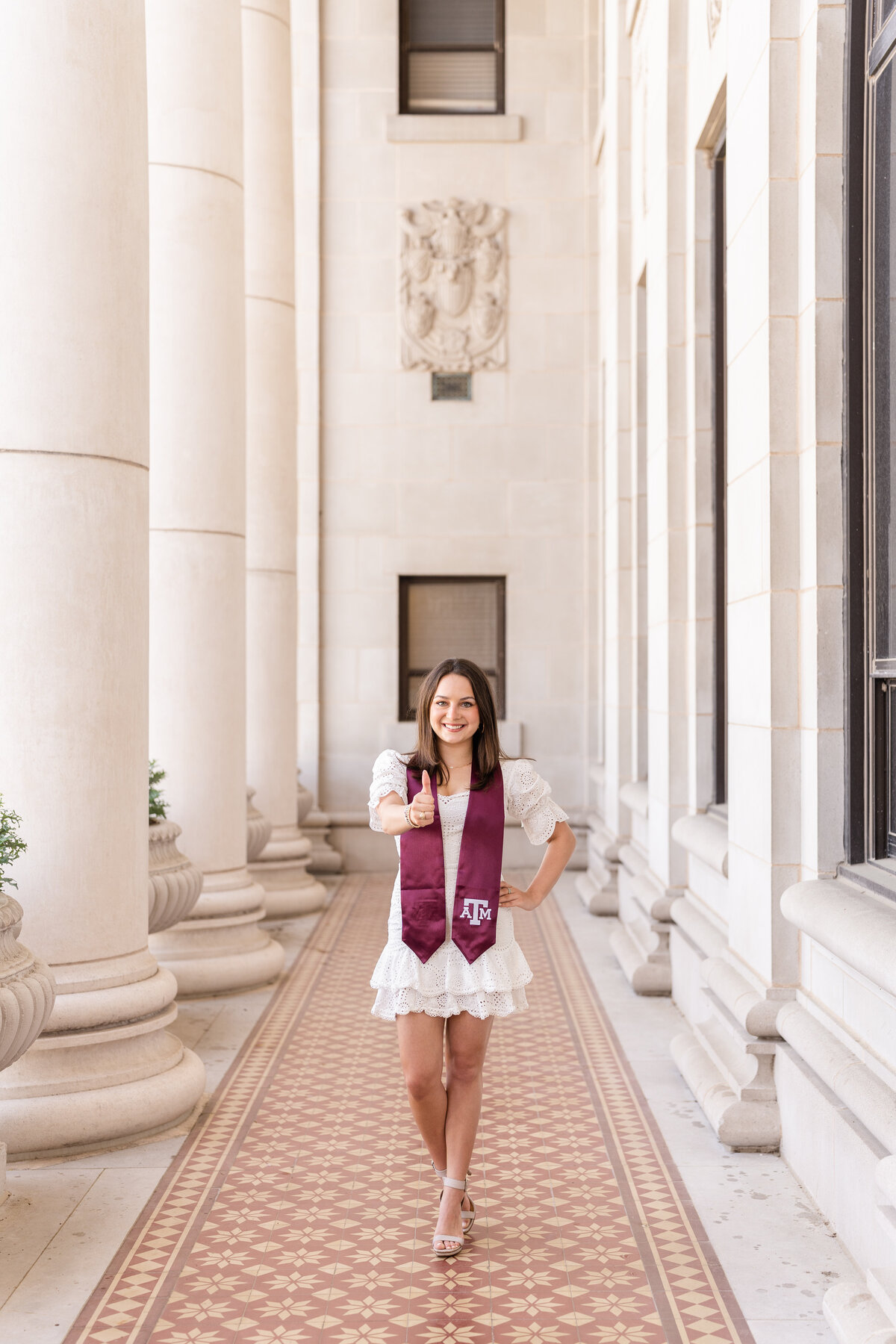 Texas A&M senior girl standing with hand on hip and thumbs up  while wearing white dress and Aggie stole in the columns of the Administration Building