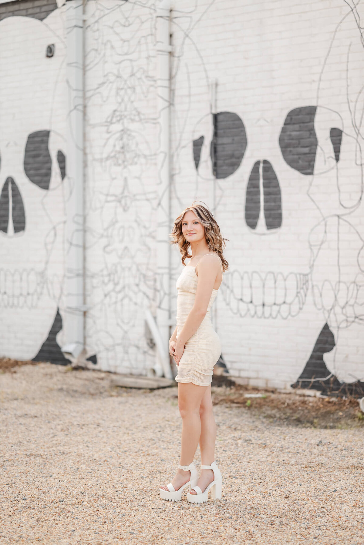 A high school senior, wearing an off-white bodycon dress and shoes, looks over her should. She is standing in front of a brick wall painted with a skull mural.