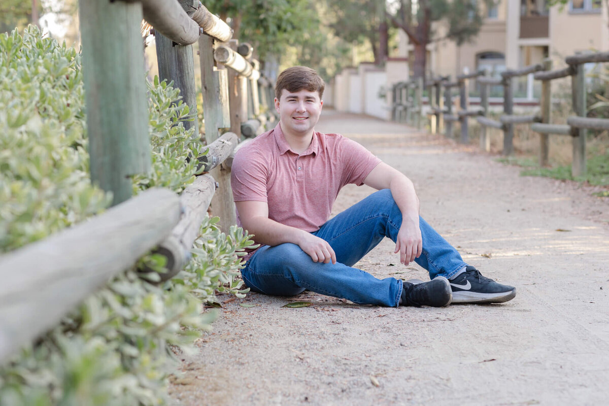 A young man in a red shirt and blue jeans, with dirty blonde hair, sitting in a gravel walking path at a park, facing the camera.