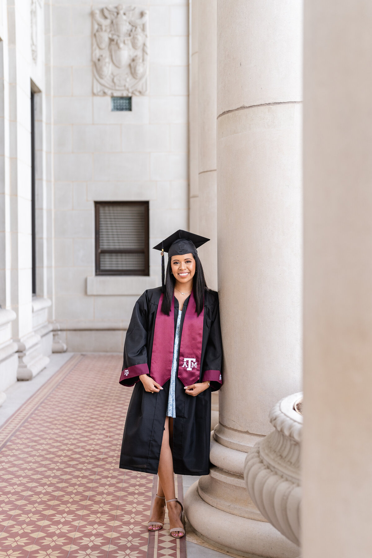 Texas A&M senior girl wearing gown, Aggie stole and cap while holding stole and leaning against column and smiling at the Administration Building