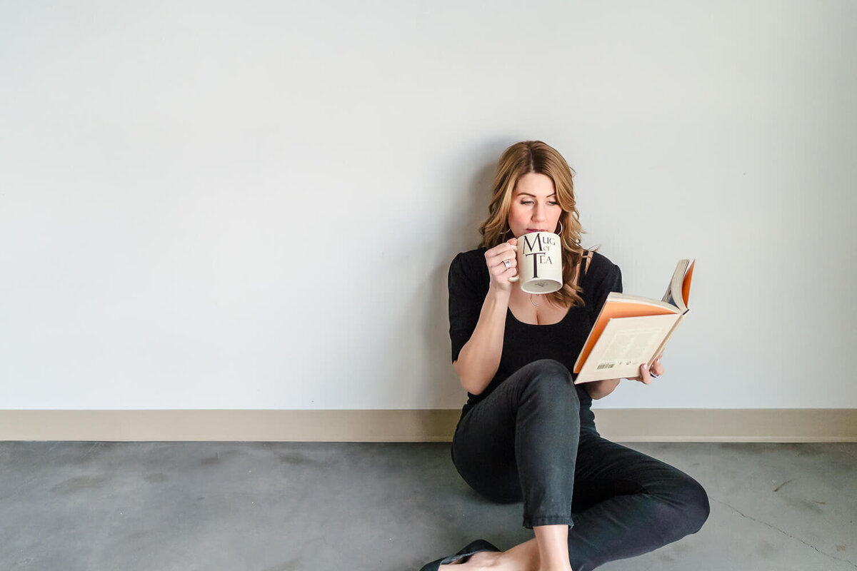 Woman sitting on floor leaning against white wall reading a book indoors and drinking tea