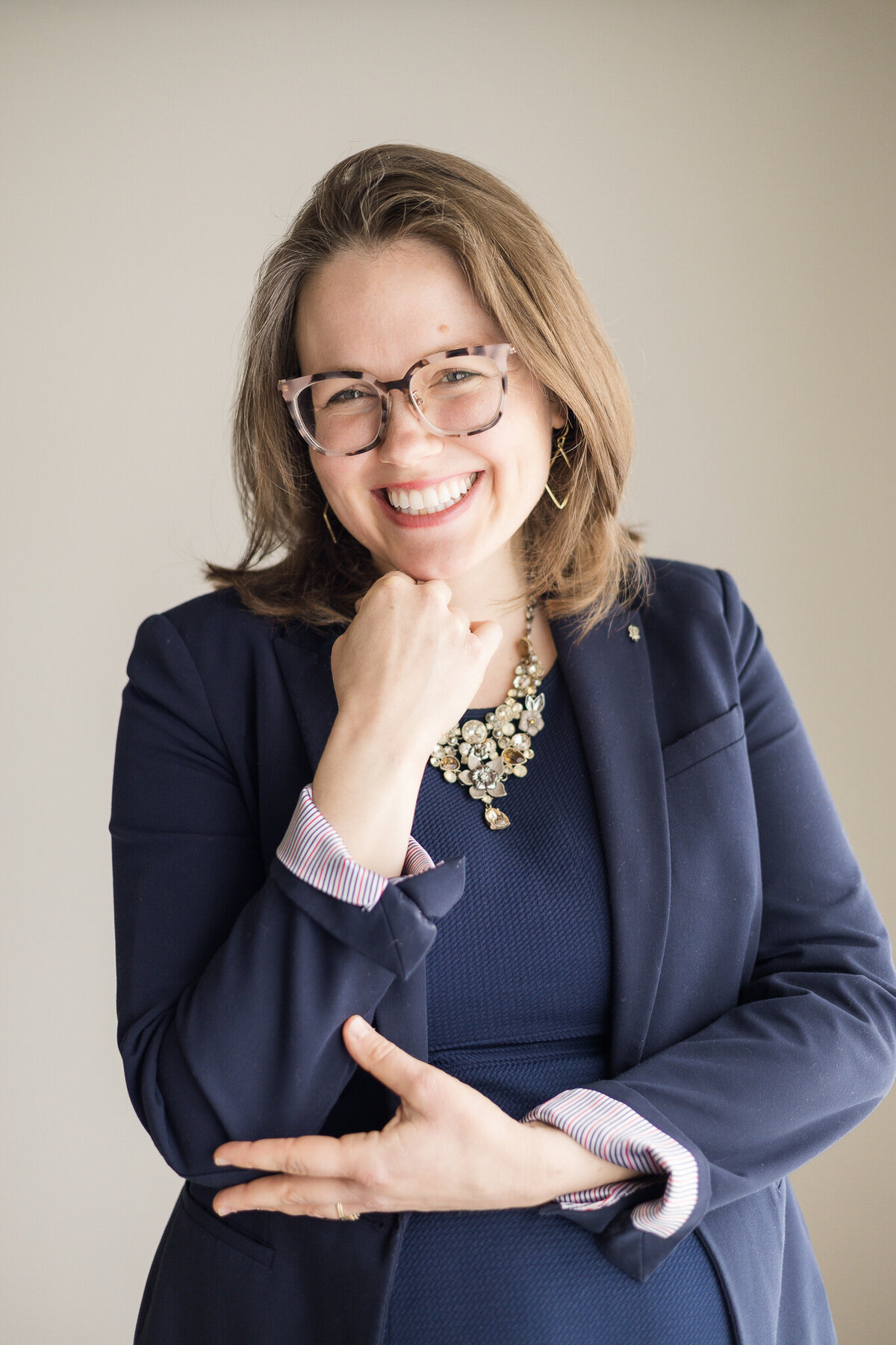 Brunette blonde pregnant female standing with her hand under her chin, wearing glasses, a dark blue dress with a long sleeve dark blue jacket, smiling at the camera