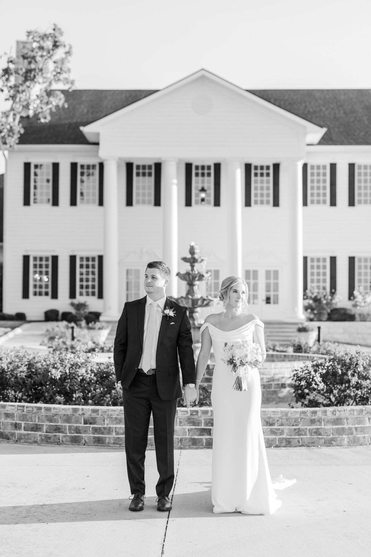 A bride and groom hold hands outside a grand white-columned estate, captured in an elegant black and white style. This editorial wedding photo highlights light and airy fine art photography, perfect for couples seeking romantic wedding portraits in the greater DFW area, including Plano, Frisco, and Highland Village.