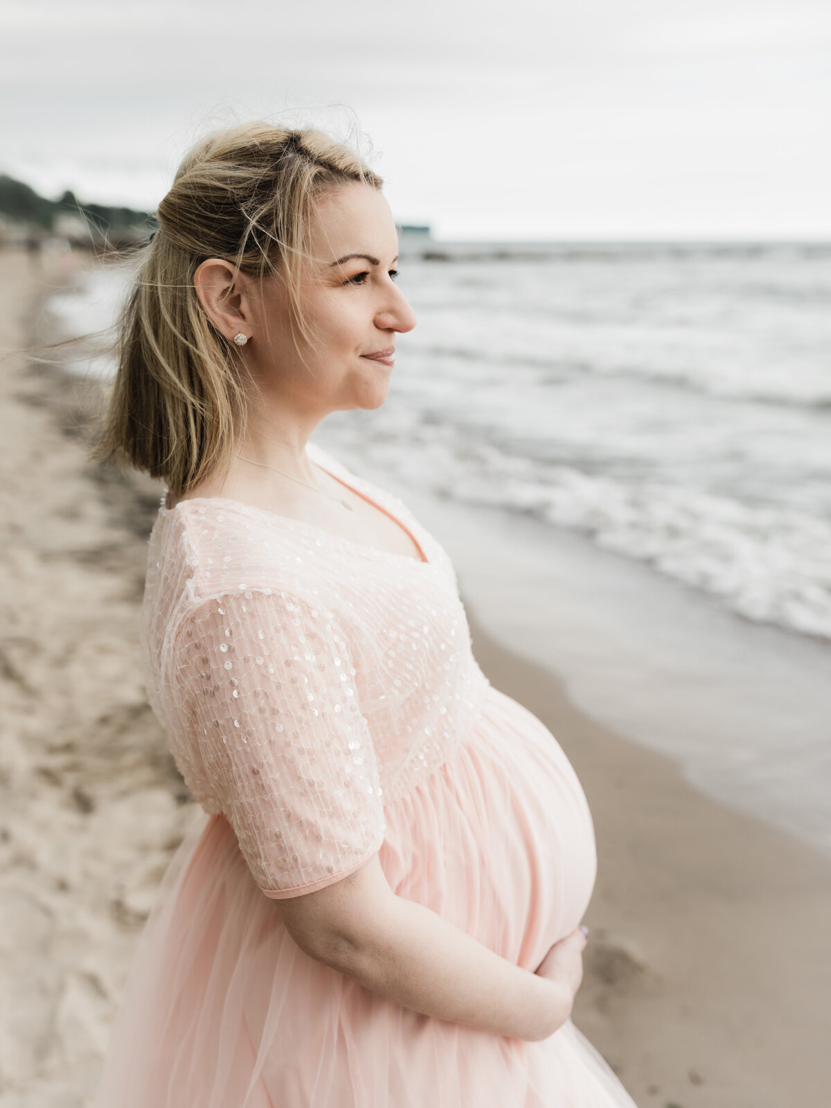 pregnant mother in pink maternity gown holding baby bump on beach for maternity photoshoot cleveland maternity photographer