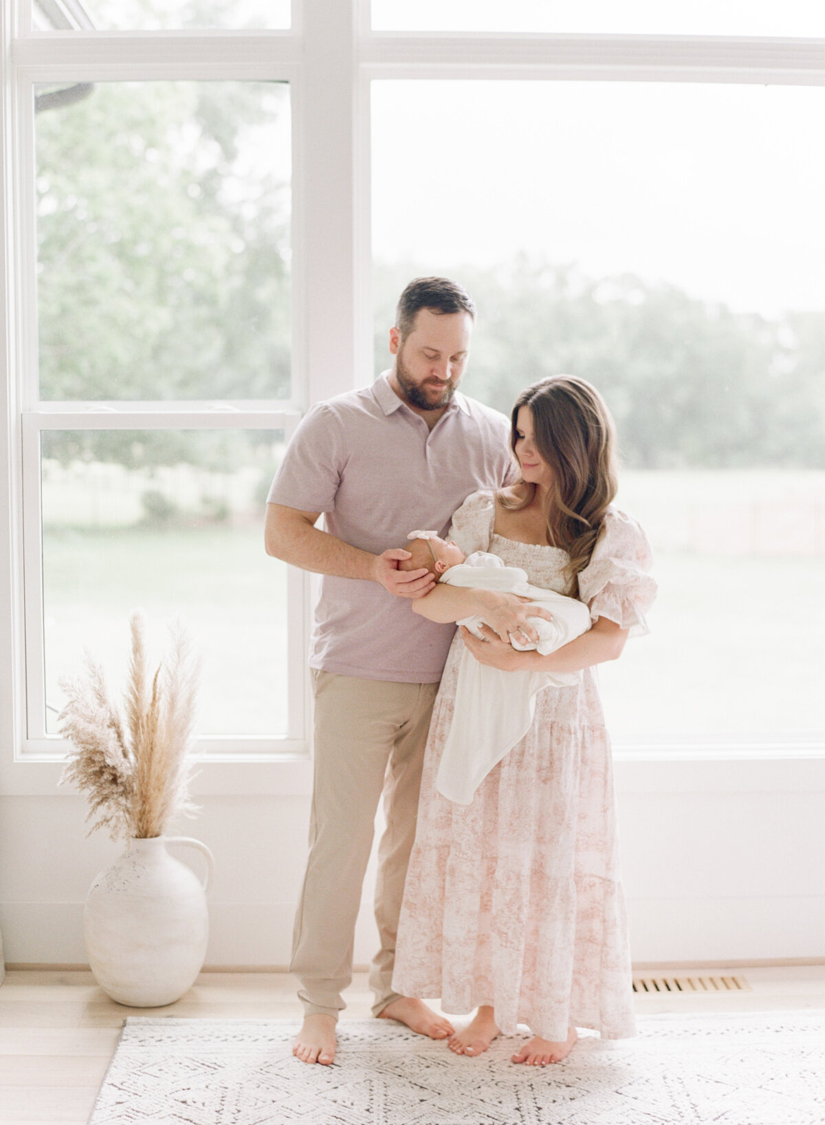 New parents stand and hold their daughter in front of large windows during their newborn session in Raleigh. Photographed by Raleigh Newborn Photographer A.J. Dunlap Photography.