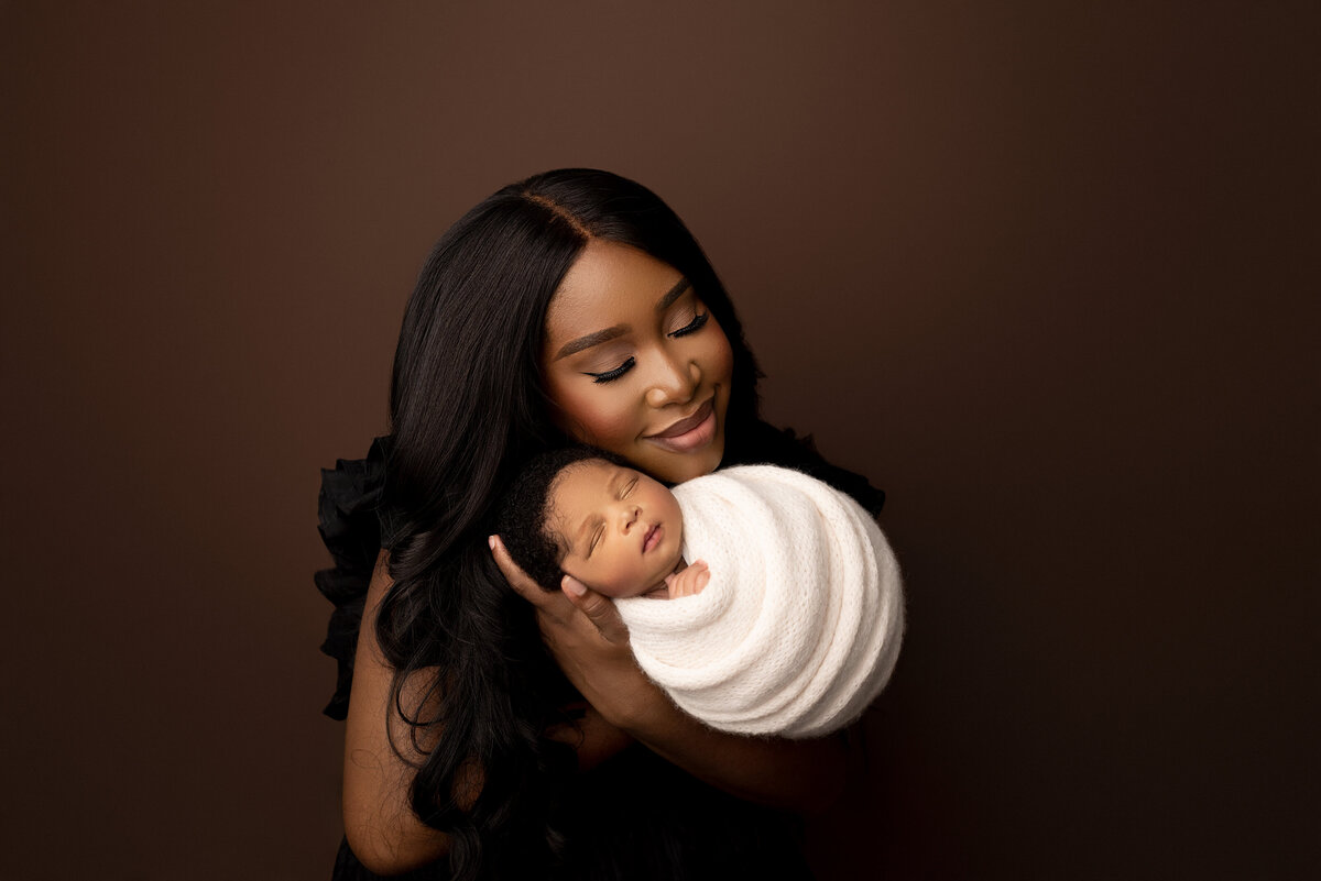 Fine art newborn baby photos captured by the best Main Line newborn baby photographer Katie Marshall. Black mom with long softly curled hair holds her newborn baby boy swaddled in white. Baby is facing the camera and sleeping. Mom is resting her cheek against baby's temple and her eyes are gently closed. She is wearing a black gown against a brown backdrop. Close-up crop chest-up.