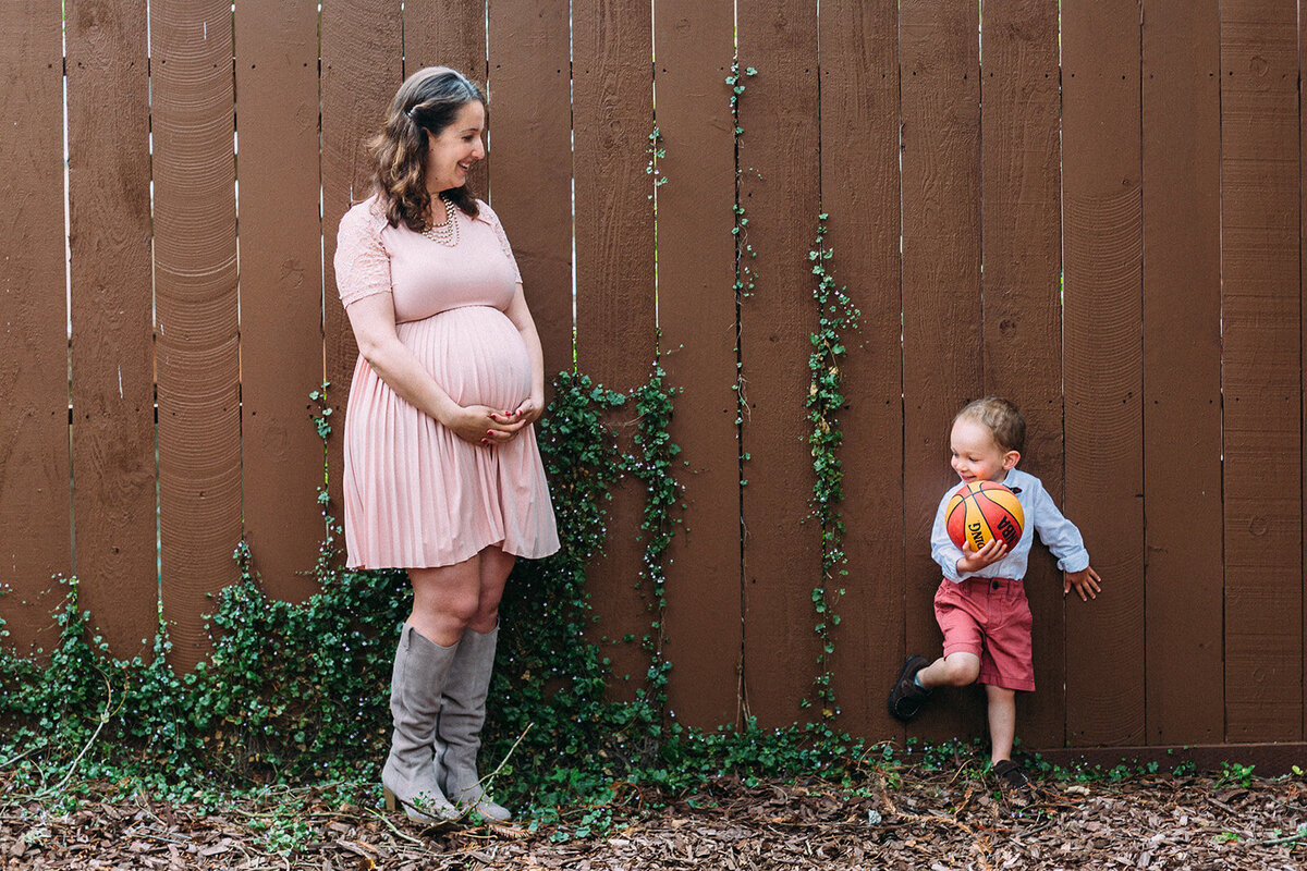 Maternity Photographer, a little child leans on a yard fence as his pregnant mother admires him