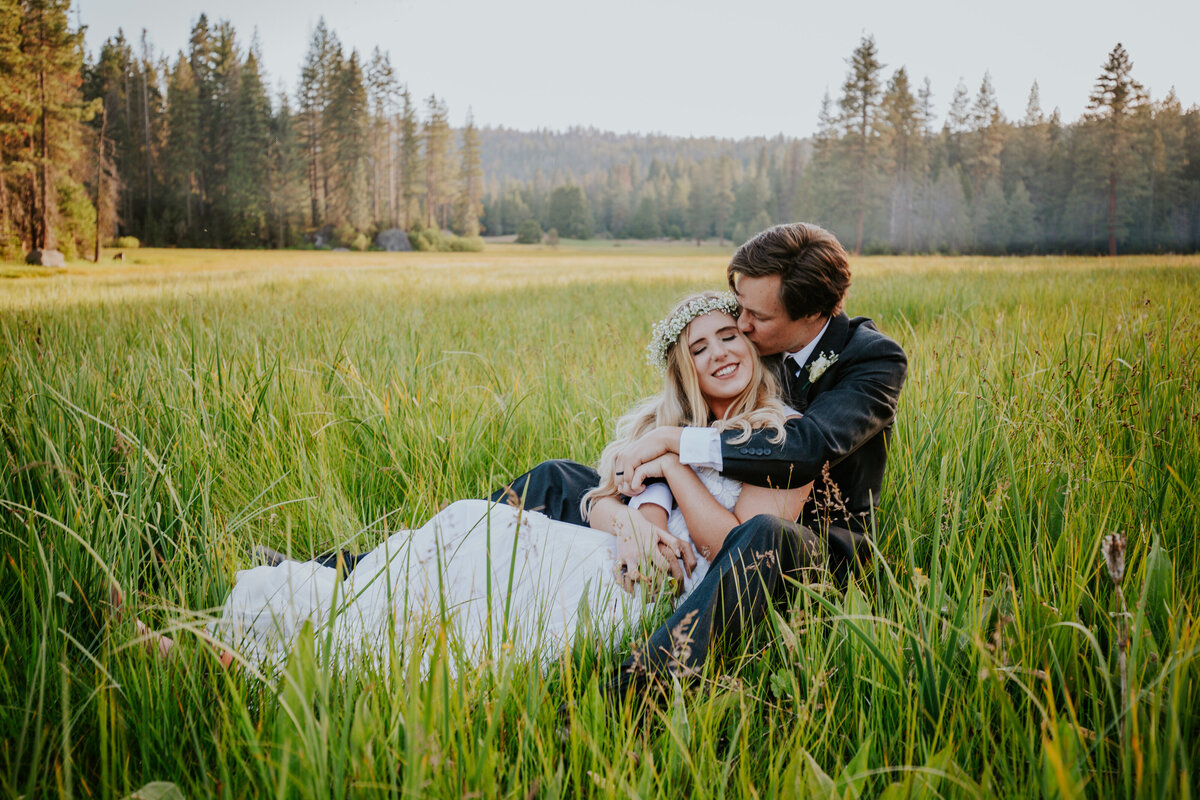 Bride leans into grooms lap as he kisses her cheek in the middle of a forest meadow.