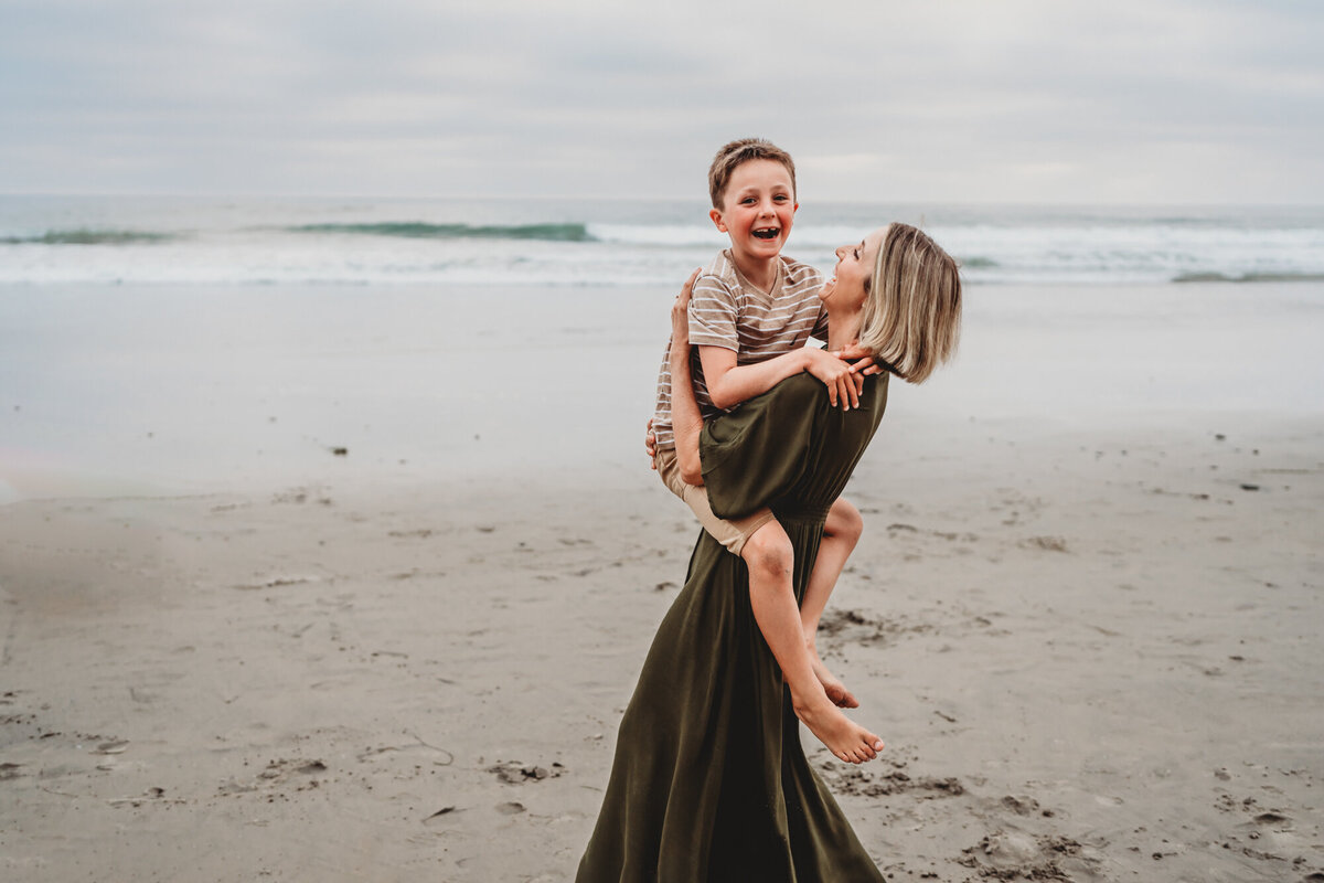 Mother laughs as she carries her delighted son on the beach during a San Diego family photo session in Del Mar.