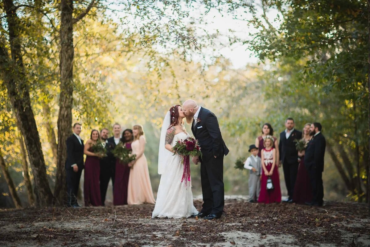 A bride and groom share a secluded moment in the woods, with their wedding party in the background