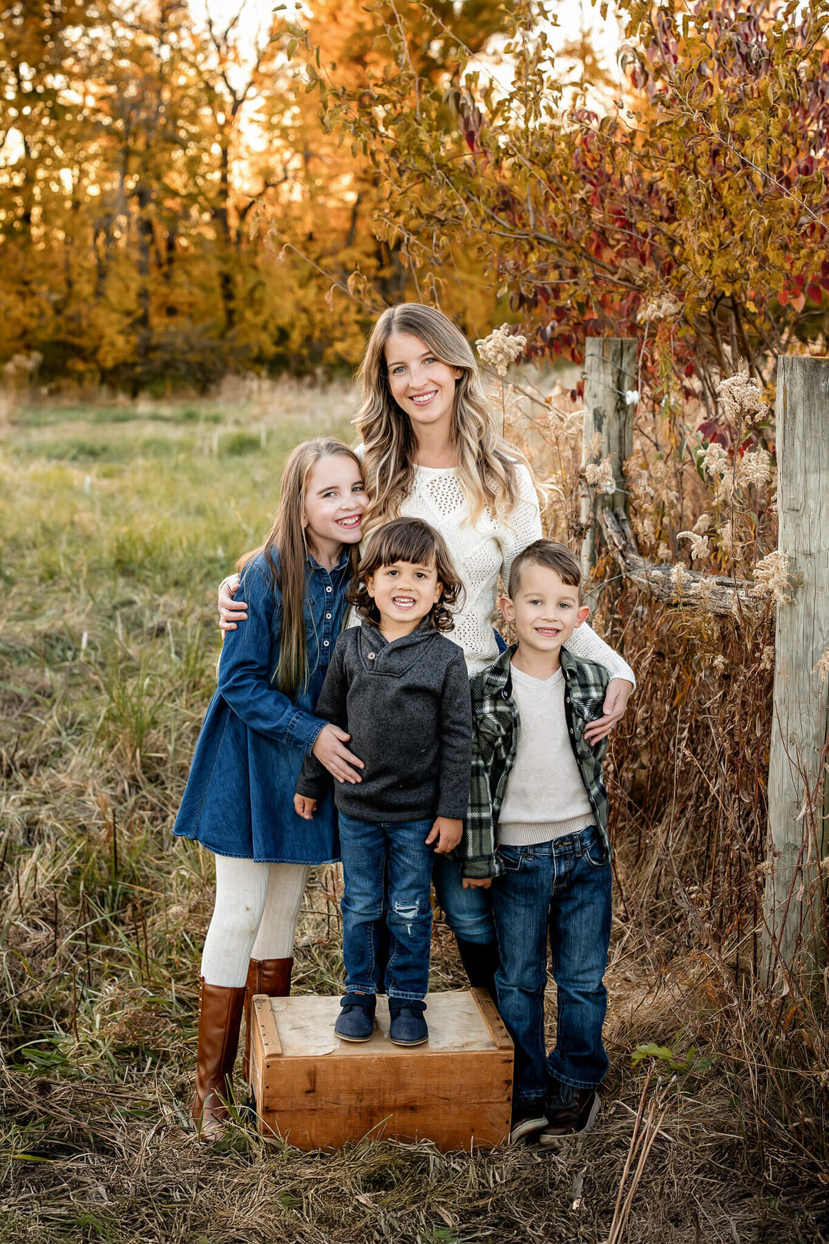 A woman and three children pose outdoors in autumn, capturing incredible moments. The woman smiles warmly behind the children—the eldest girl dons a blue jacket on the left, while two young boys stand joyfully on a wooden box amidst vibrant fall foliage.