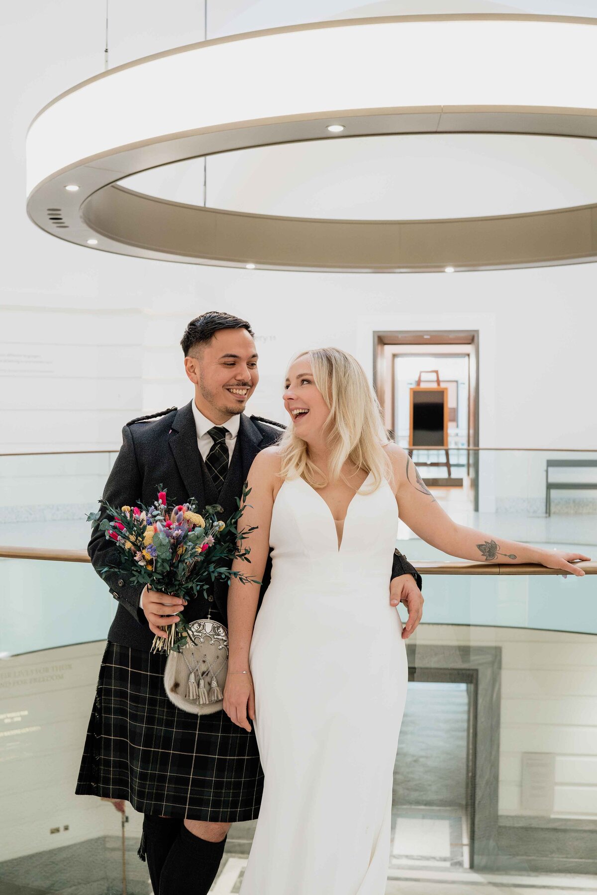 A couple stands on a modern balcony in Aberdeen Art Gallery, captured by an Aberdeen wedding photographer. The man wears a traditional kilt and holds a bouquet, while the woman is in a white wedding dress; they smile at each other amidst circular lighting and contemporary architecture.