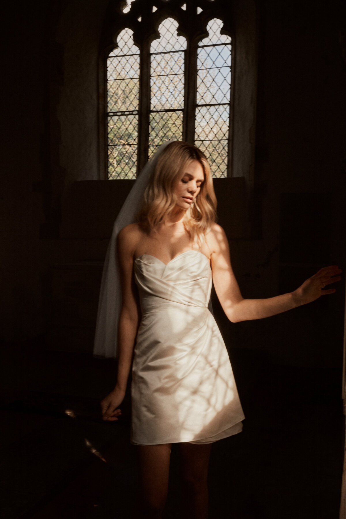 Bride with sleeveless mini wrap wedding dress in pure silk crepe de chine, stood in church window