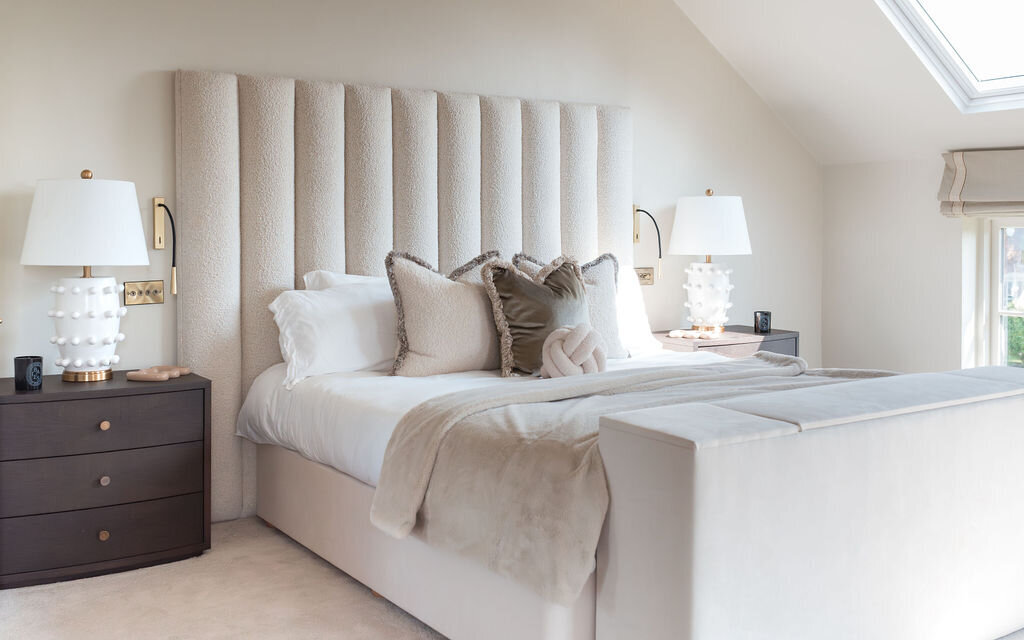 A well-lit bedroom featuring a large plush bed with a tall, tufted headboard. The bed is flanked by two dark wooden nightstands, each with a white lamp. The room has a neutral color palette with white and beige bedding and some accent pillows.