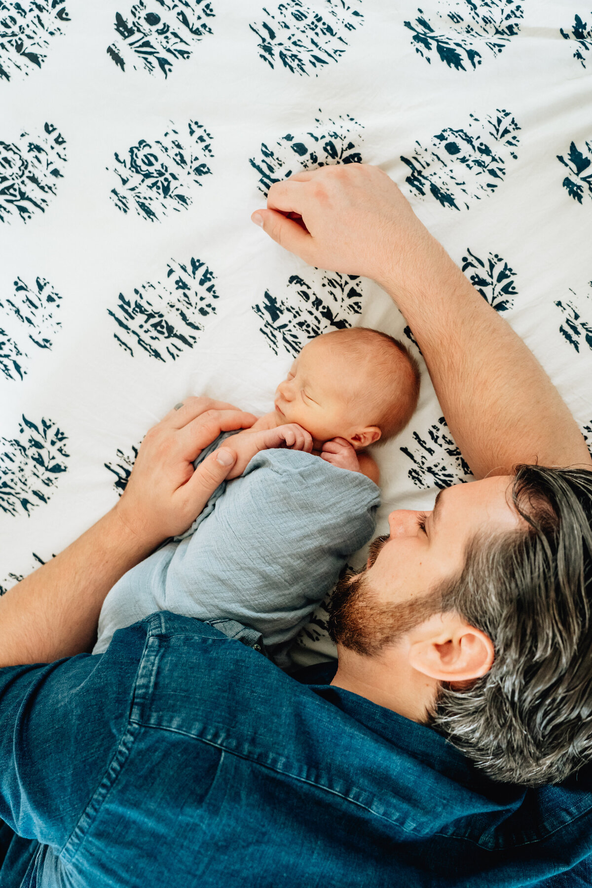 A father lying on a bed next to his newborn baby, who is swaddled in a blue blanket.