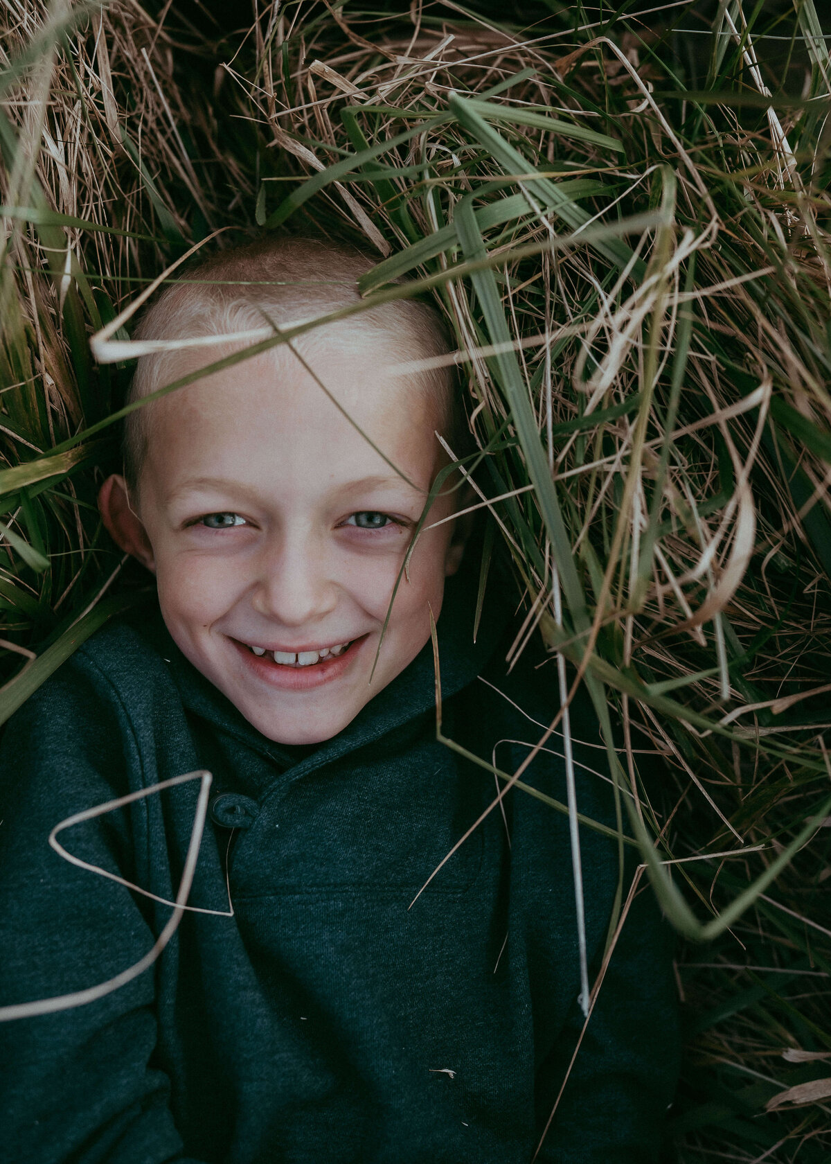 A young fair skin blonde hair boy grins at the camera while wearing a navy blue shirt and being enveloped by the tall grass as he's laying in.
