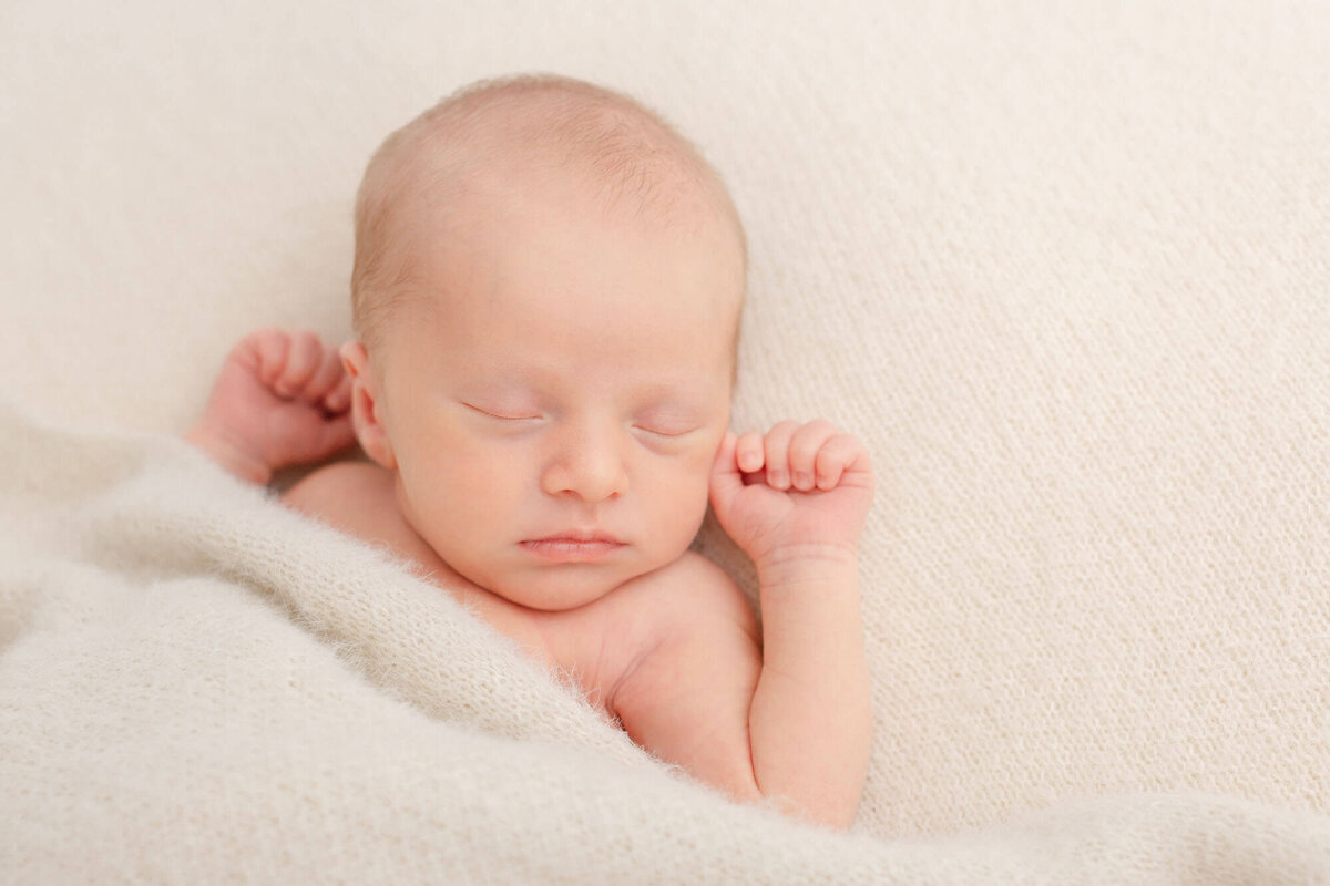 Baby sleeping with his little hands up in fists by his head. He is sleeping on a beige blanket and has a cozy little beige blanket pulled up to his chin. He is sleeping peacefully.